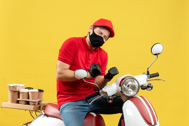 Vista superior del mensajero feliz con blusa roja y guantes de sombrero en máscara médica entregando orden sentado en scooter