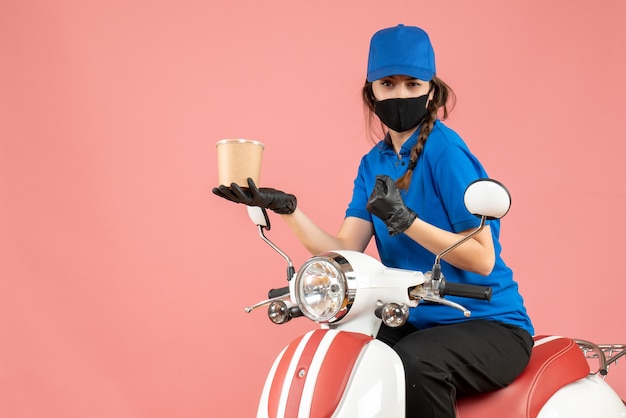 Vista superior del mensajero femenino confiado con máscara médica y guantes sentado en scooter entregando pedidos sobre fondo melocotón pastel