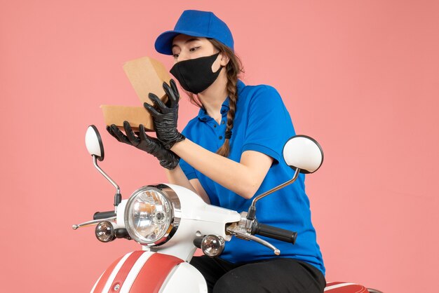 Vista superior del mensajero femenino enfocado joven con máscara médica y guantes abriendo caja en melocotón pastel