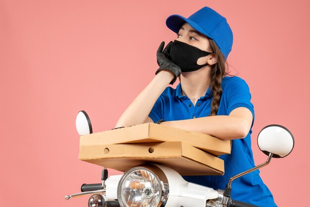 Vista superior del mensajero femenino joven pensante con máscara médica y guantes sosteniendo cajas en melocotón pastel