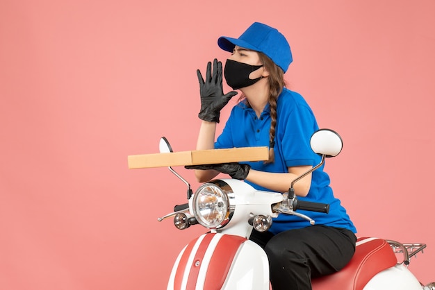 Vista superior del mensajero femenino con máscara médica y guantes sentado en scooter entregando pedidos llamando a otros sobre fondo melocotón pastel