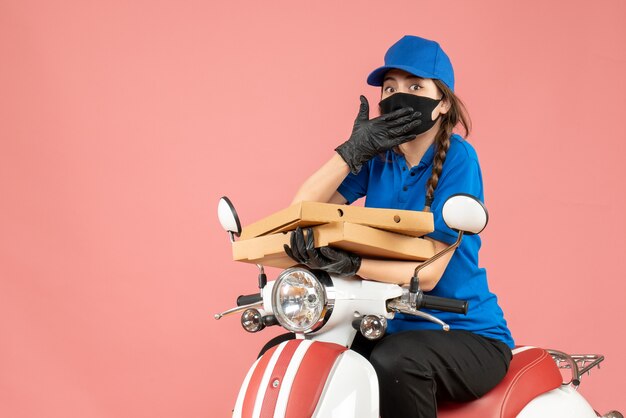 Vista superior del mensajero femenino preocupado joven con máscara médica y guantes sentado en scooter entregando pedidos en melocotón pastel