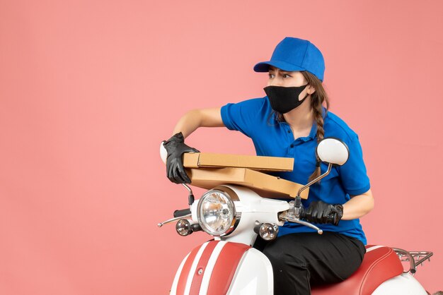 Vista superior del mensajero femenino preocupado con máscara médica y guantes sentado en scooter entregando pedidos en melocotón pastel