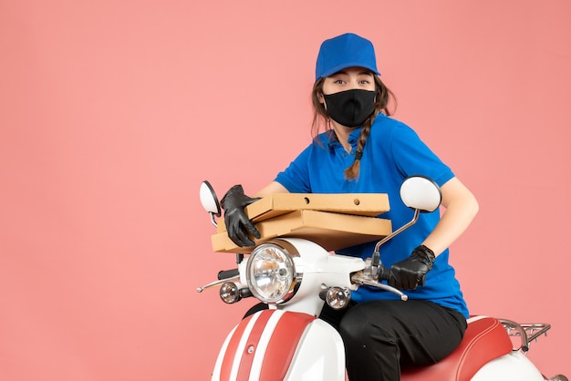 Foto gratuita vista superior del mensajero femenino sonriente joven con máscara médica y guantes sentado en scooter entregando pedidos en melocotón pastel