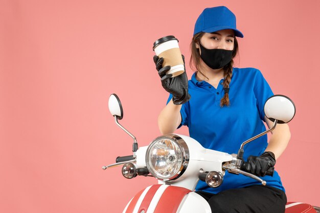 Vista superior del mensajero femenino trabajador con máscara médica negra y guantes entregando pedidos sobre fondo melocotón