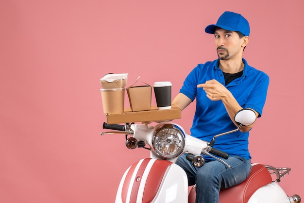 Vista superior del mensajero sorprendido con sombrero sentado en scooter apuntando hacia adelante en melocotón pastel