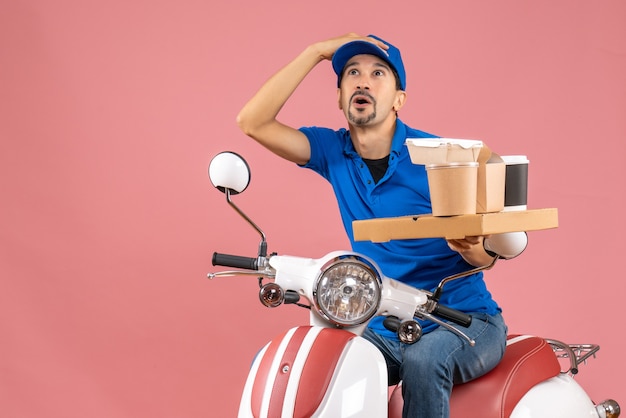 Vista superior del mensajero sorprendido con sombrero sentado en scooter mirando hacia arriba en melocotón pastel