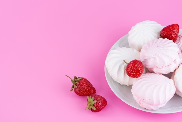 Una vista superior de merengues blancos dentro de la placa junto con fresas rojas frescas en el escritorio rosa, azúcar de galleta de frutas y bayas