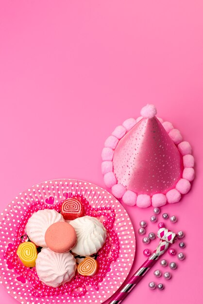 Una vista superior de merengues y mermeladas dentro de la placa junto con silbatos de cumpleaños y gorro de cumpleaños en rosa, color dulce de azúcar de malvavisco