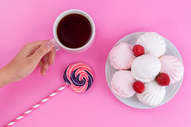 Una vista superior de merengues y té hembra sosteniendo una taza de té junto con una piruleta en rosa, galleta de pastel de té
