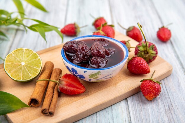 Vista superior de mermelada de fresa en un recipiente sobre una tabla de cocina de madera con fresas frescas con canela en rama con limón sobre un fondo de madera gris