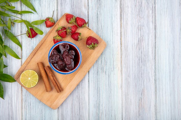 Vista superior de mermelada de fresa en un recipiente sobre una tabla de cocina de madera con ramas de canela sobre un fondo de madera gris con espacio de copia