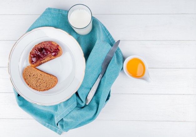 Vista superior de mermelada de fresa untada en rodajas de pan de centeno en un plato con un cuchillo de leche sobre tela azul y mantequilla derretida en la mesa de madera con espacio de copia