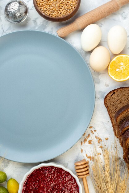 Vista superior de la mesa de desayuno con placa azul, huevos, mermelada, gelatina, pan oscuro sobre fondo blanco, masa de café, pastel, horno, té de la mañana, bollos de huevo