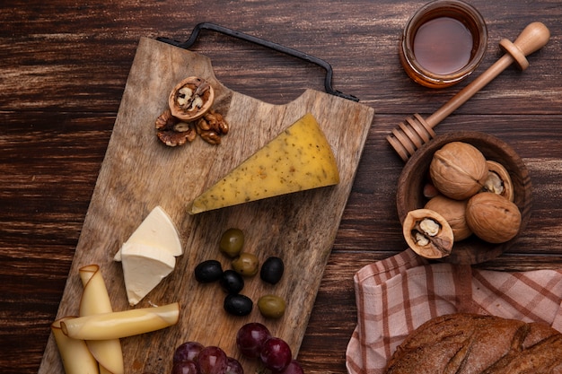 Vista superior de la miel en un tarro con nueces y una hogaza de pan negro con variedades de quesos y uvas en un soporte sobre un fondo de madera