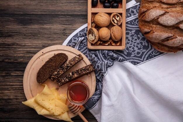 Vista superior de la miel en un tarro con pan negro y queso en un soporte con nueces sobre un fondo de madera