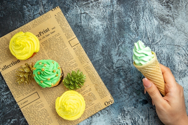 Vista superior mini cupcakes adornos navideños en helado de periódico en mano femenina sobre superficie oscura