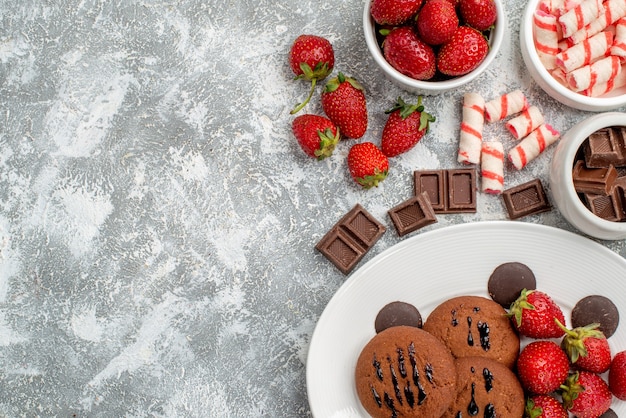 Foto gratuita vista superior de la mitad de las galletas, fresas y bombones redondos en la placa ovalada blanca y cuencos de dulces, fresas, chocolates en la mesa gris-blanca