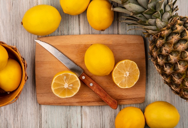 Vista superior de la mitad de los limones frescos en una tabla de cocina de madera con un cuchillo con limones en un cubo con limones y piña aislado en una superficie de madera gris