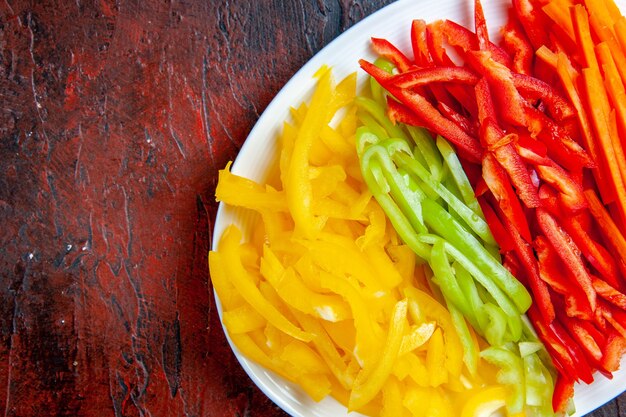 Vista superior de la mitad de los pimientos cortados de colores en un plato blanco sobre la mesa rojo oscuro