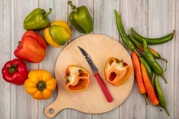 Vista superior de la mitad de pimientos naranjas en una tabla de cocina de madera con un cuchillo con pimientos de colores aislados sobre un fondo de madera gris