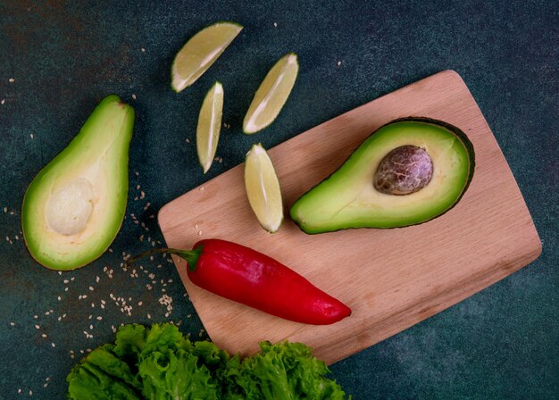 Vista superior mitades de aguacate en una pizarra con pimiento rojo limón y lechuga sobre un fondo verde oscuro