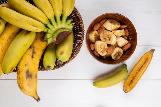 Vista superior de un montón de fruta de plátano en una cesta de mimbre y un tazón con rodajas de plátano y almendras en blanco