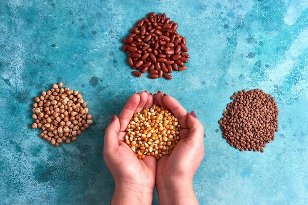 Vista superior de montones de garbanzos, frijoles rojos y lentejas marrones con la mano sosteniendo los granos de maíz