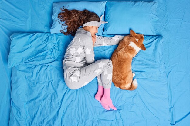 Vista superior de la morena joven europea en pijama duerme junto con su mascota favorita ve dulces sueños se siente cómodo tiene poses saludables para dormir en la cama. Personas relajación animales concepto de hora de acostarse