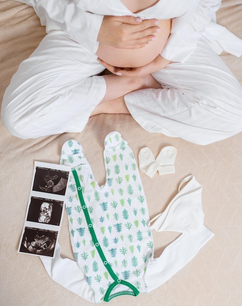 Vista superior de una mujer anónima con un pijama blanco y acogedor sentada en la cama con una imagen de ultrasonido de un traje de cuerpo y calcetines lindos en el útero mientras acaricia el vientre embarazado y espera un bebé y parte de la familia