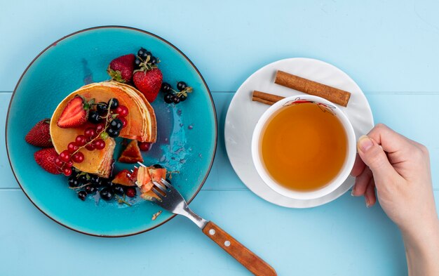 Vista superior mujer bebe taza de té con panqueques con fresas grosellas rojas y negras sobre una mesa azul