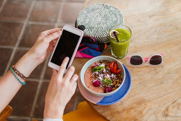 Vista superior de una mujer bonita en pantalones amarillos usando el teléfono y disfrutando de sabrosa comida vegana