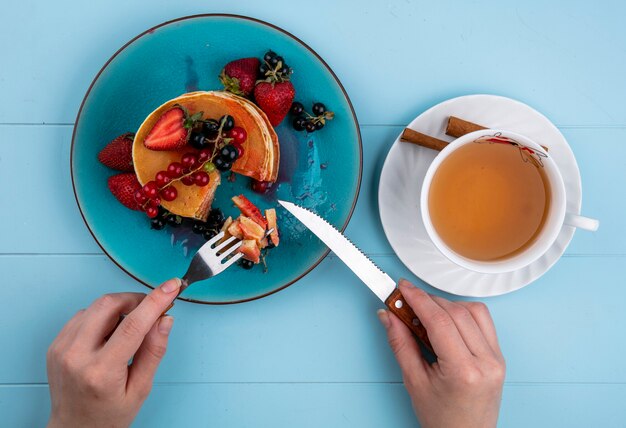 Vista superior mujer come panqueques con fresas grosellas rojas y negras y una taza de té en una mesa azul