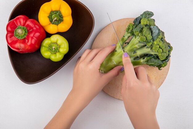 Vista superior mujer corta brócoli sobre una tabla de cortar con pimientos de colores en un recipiente sobre un fondo blanco.