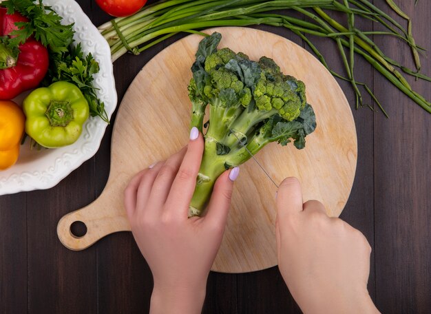 Vista superior mujer corta brócoli en tabla de cortar con pimientos y cebollas verdes sobre fondo de madera