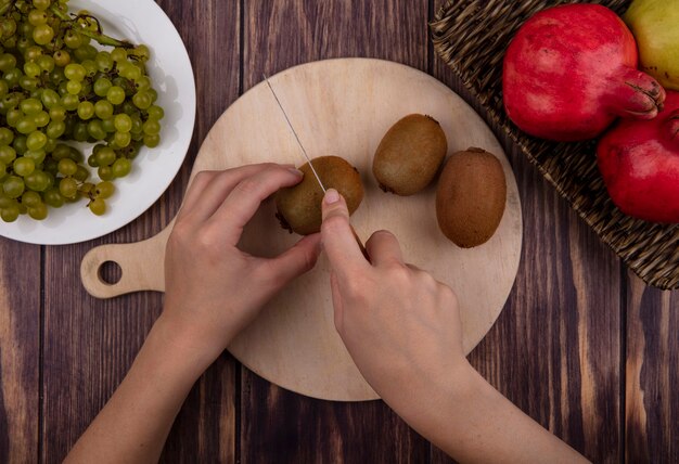 Vista superior mujer corta kiwi en tabla de cortar con uvas y granadas en pared de madera