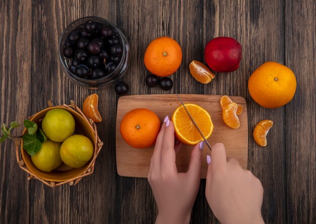 Vista superior mujer corta naranja sobre una tabla de cortar con ciruelas en una canasta y ciruelas cereza en un frasco sobre fondo de madera