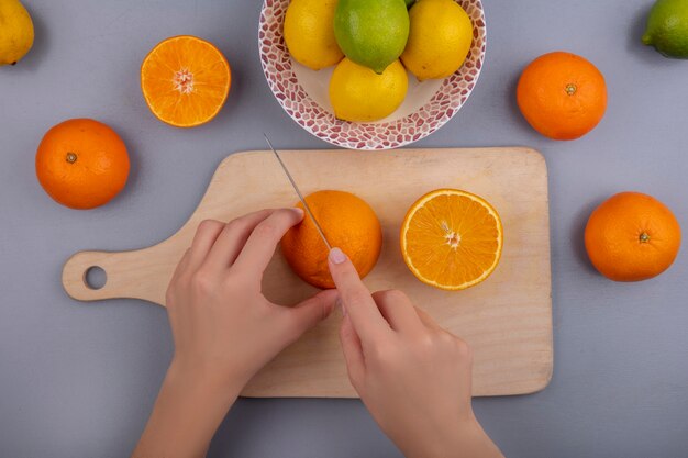 Vista superior mujer corta naranjas en tabla de cortar con limones y limas en placa sobre fondo gris