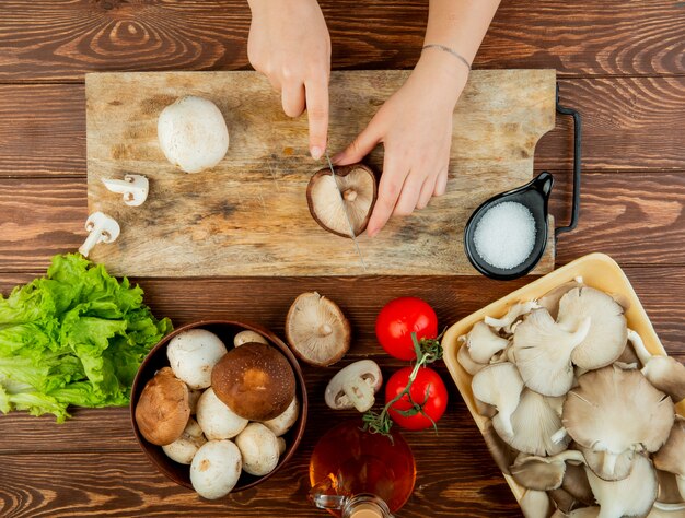 Vista superior de una mujer cortando champiñones frescos en una tabla para cortar madera y tomates con lechuga en madera rústica