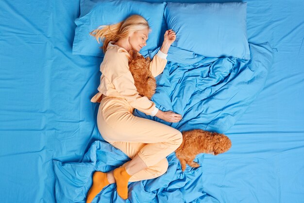 La vista superior de la mujer dormida relajada tiene una siesta saludable en la cama posa con dos cachorros vestidos con ropa de dormir que disfruta de la comodidad en ropa de cama suave y ve dulces sueños. Amistad entre personas y animales.