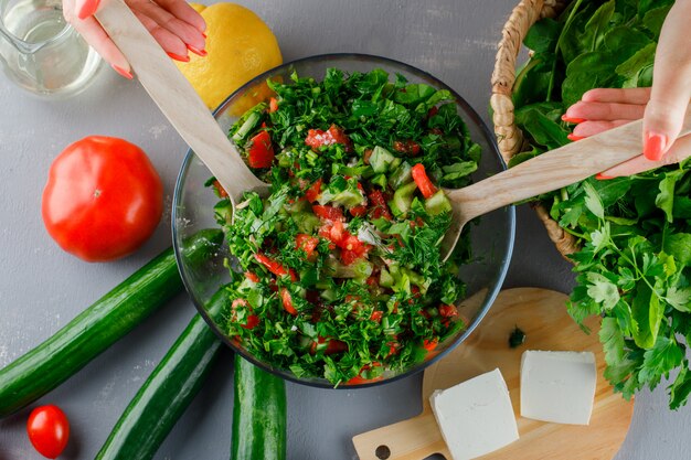 Vista superior mujer haciendo ensalada de verduras en un recipiente de vidrio con tomates, queso, verduras, pepino en superficie gris