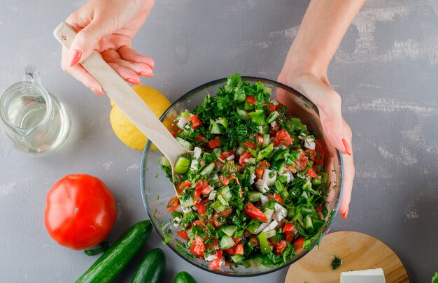 Vista superior mujer haciendo ensalada de verduras en un tazón de vidrio con tomate, pepino, limón sobre superficie gris