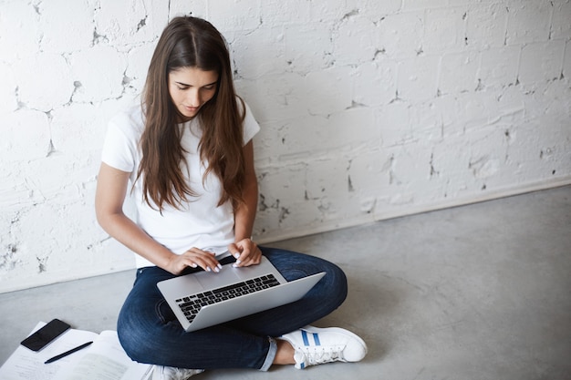 Foto gratuita vista superior de la mujer joven creativa, usando la computadora portátil mientras está sentado en el piso, apoyado en la pared, rodeado de aparatos y libros. la diseñadora trabaja en un nuevo proyecto que necesita presentar mañana. concepto de estudio