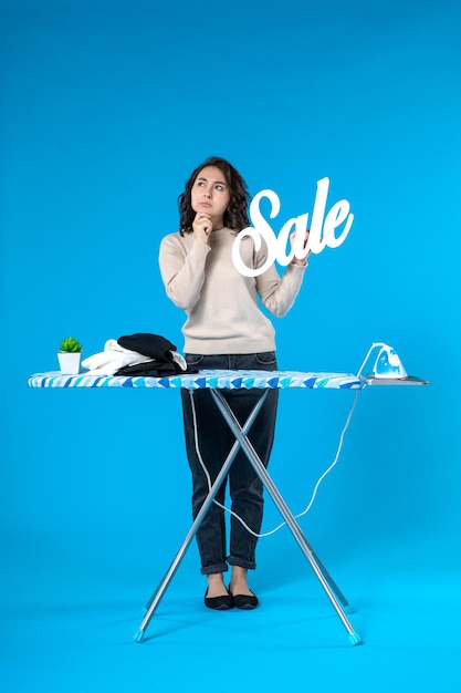 Vista superior de la mujer joven pensativa de pie detrás de la tabla de planchar y mostrando el icono de venta sobre fondo azul.