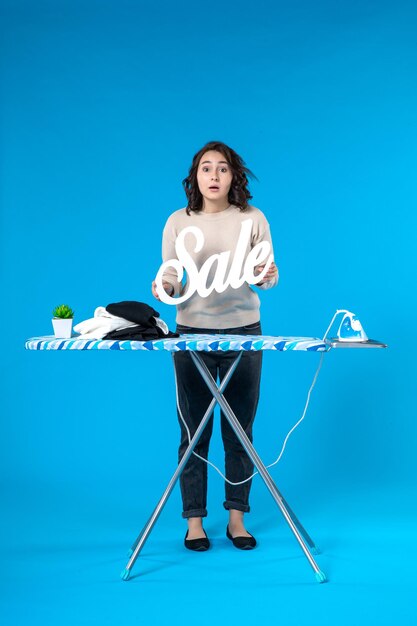 Vista superior de la mujer joven sorprendida de pie detrás de la tabla de planchar y mostrando el icono de venta en la superficie azul