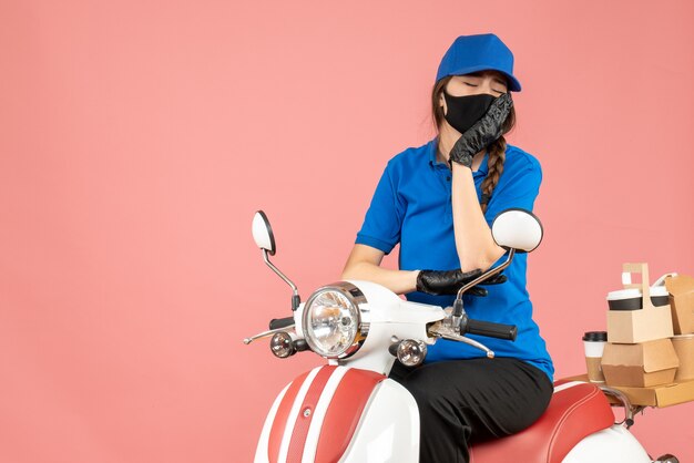 Vista superior de la mujer de mensajería cansada con máscara médica y guantes sentado en scooter entregando pedidos en melocotón pastel