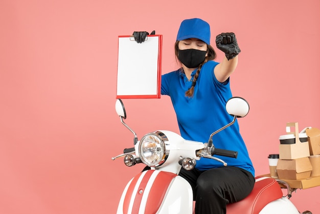 Vista superior de la mujer de mensajería feliz con máscara médica y guantes sentado en scooter sosteniendo hojas de papel vacías entregando pedidos sobre fondo melocotón pastel