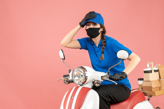 Vista superior de la mujer de mensajería sorprendida con máscara médica y guantes sentado en scooter entregando pedidos en melocotón pastel