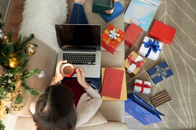 Vista superior de la mujer sentada en el sofá con laptop y café rodeada de numerosas cajas de regalo