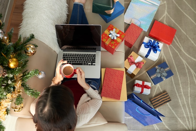 Vista superior de la mujer sentada en el sofá con laptop y café rodeada de numerosas cajas de regalo